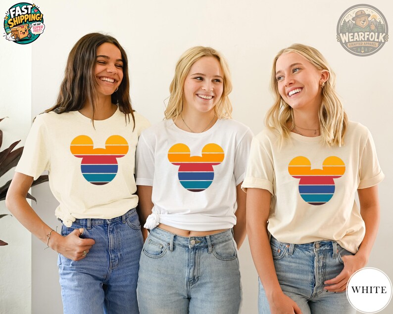 Three women wearing cream and white tees with a colorful Mickey Mouse head graphic in orange, red, and blue horizontal stripes.