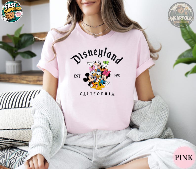 A woman wearing a light pink tee with Disneyland vintage graphic featuring Mickey, Minnie, Goofy, and Donald Duck with text reading "Disneyland EST 1955 California".