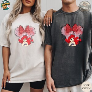 A woman and man wearing matching Bella Canvas tees with Ariel graphic featuring mouse ears with red bow and red-haired mermaid character in pink and watercolor style.