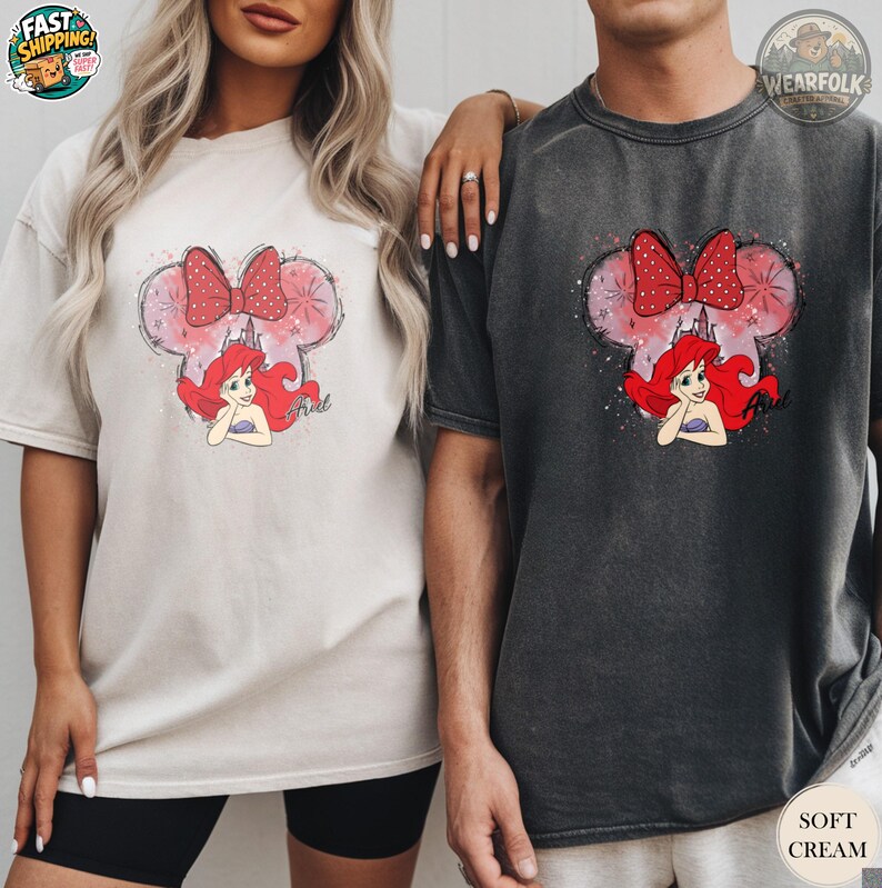 A woman and man wearing matching Bella Canvas tees with Ariel graphic featuring mouse ears with red bow and red-haired mermaid character in pink and watercolor style.