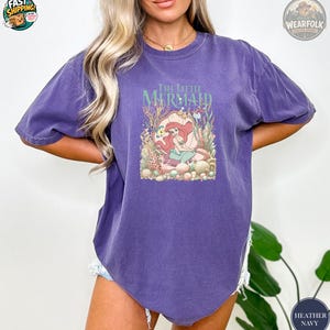 A woman wearing a purple vintage tee with The Little Mermaid graphic featuring Ariana character surrounded by colorful coral and sea creatures.