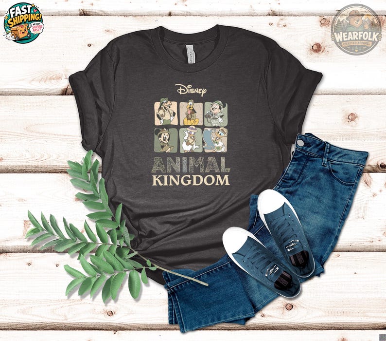 Flat lay of a dark grey t-shirt with Disney Animal Kingdom safari-themed graphic featuring Goofy, Donald, Minnie, Mickey, and Daisy Duck in explorer outfits, with text reading "Disney Animal Kingdom."
