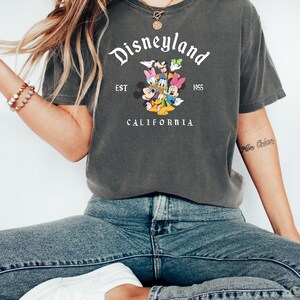 K&ouml;nnte beinhalten: Dunkelgraues T-Shirt mit dem Disneyland-Logo und den Zeichentrickfiguren Mickey, Minnie, Donald, Daisy, Goofy und Pluto. Der Text "EST 1955 CALIFORNIA" ist darunter aufgedruckt. Das T-Shirt wird mit blauen Jeans und wei&szlig;en Turnschuhen getragen.