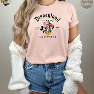 A woman wearing a pink Bella Canvas tee with Disneyland California EST 1955 graphic featuring Mickey Mouse and classic Disney characters.