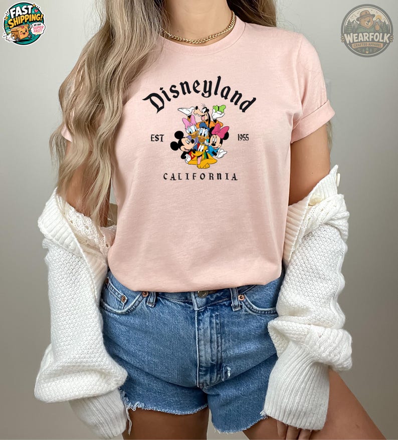 A woman wearing a pink Bella Canvas tee with Disneyland California EST 1955 graphic featuring Mickey Mouse and classic Disney characters.