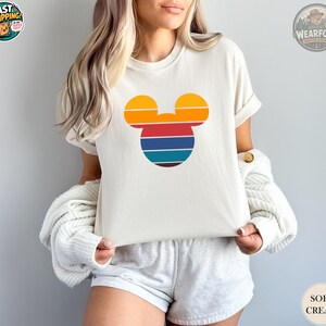 May include: Cream-colored t-shirt featuring a retro-style Mickey Mouse head design in orange, yellow, red, blue, and brown stripes. The shirt has short sleeves and a relaxed fit. The words "SOFT CREAM" are printed on the bottom right.
