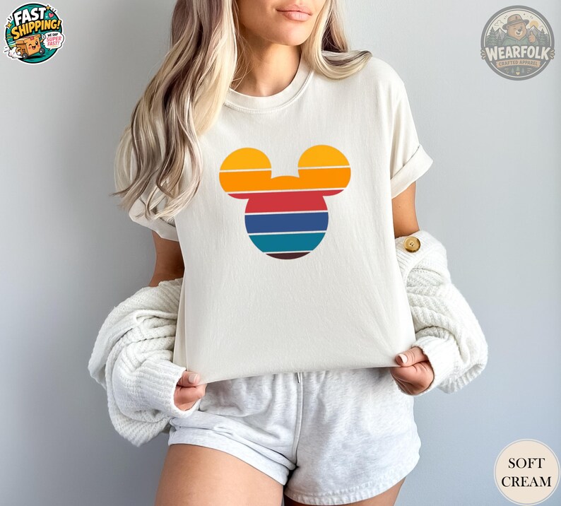 May include: Cream-colored t-shirt featuring a retro-style Mickey Mouse head design in orange, yellow, red, blue, and brown stripes. The shirt has short sleeves and a relaxed fit. The words "SOFT CREAM" are printed on the bottom right.