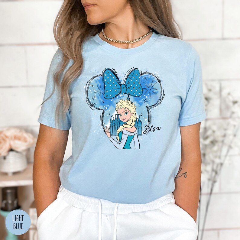 A woman wearing a light blue tee with Elsa from Frozen inside a Mickey Mouse head silhouette with a blue polka dot bow and castle graphic.