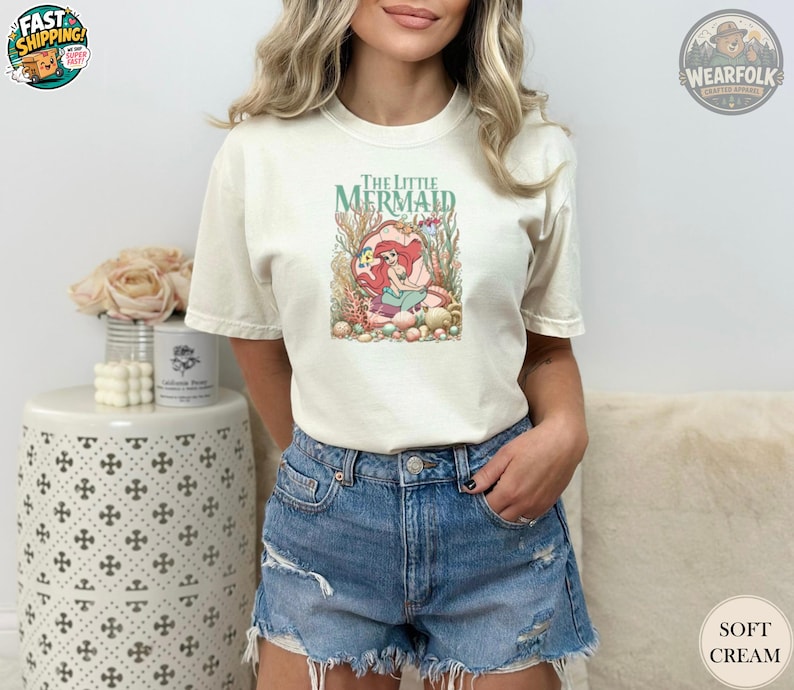 Woman wearing a soft cream tee featuring The Little Mermaid graphic with Ariana and colorful underwater coral and shells design.