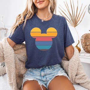 A woman wearing a heather navy tee with a colorful striped Mickey Mouse head graphic in yellow, red, blue, and teal.