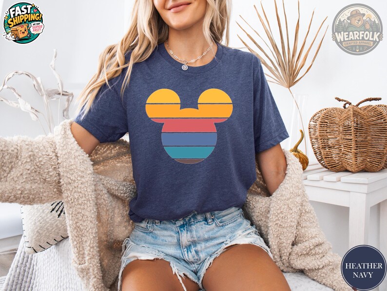 A woman wearing a heather navy tee with a colorful striped Mickey Mouse head graphic in yellow, red, blue, and teal.