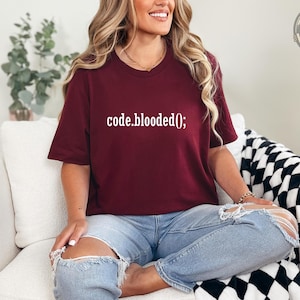 May include: A maroon short-sleeved t-shirt featuring the white text code.blooded(); printed across the chest. The shirt has a crew neckline and a standard fit, paired with light blue distressed denim jeans that have large rips at the knees.