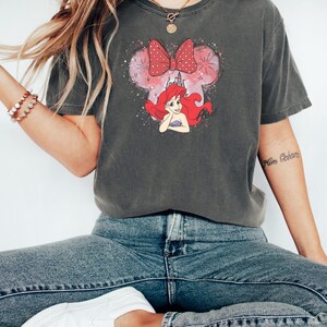 Woman wearing a charcoal gray Bella Canvas tee with Ariel graphic featuring her red hair, pink bow, and watercolor mouse ears design.