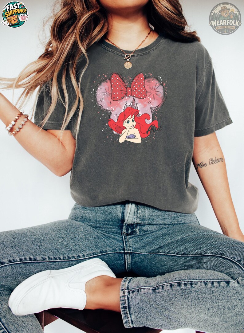 Woman wearing a charcoal gray Bella Canvas tee with Ariel graphic featuring her red hair, pink bow, and watercolor mouse ears design.