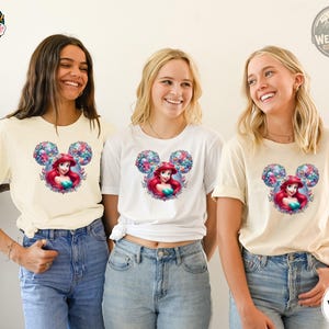Three women wearing cream and white tees with a Disney Princess Ariel graphic surrounded by colorful floral Mickey ears design.
