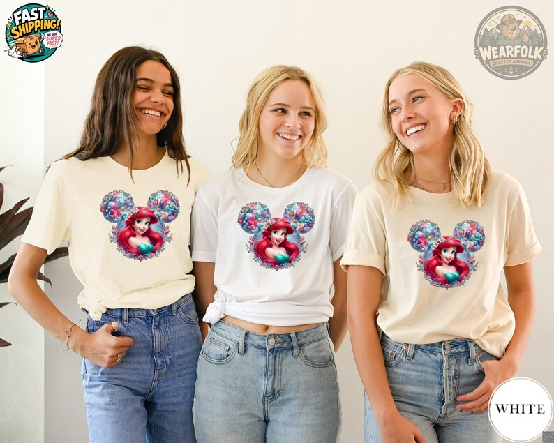Three women wearing cream and white tees with a Disney Princess Ariel graphic surrounded by colorful floral Mickey ears design.
