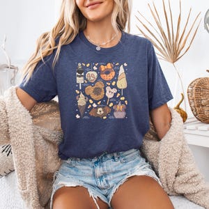 A woman wearing a heather navy tee with an illustrated baking-themed graphic featuring cookies, bread, pastries, and kitchen items with text labels.