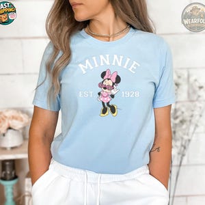 A woman wearing a light blue t-shirt featuring Minnie Mouse in sunglasses and a pink polka dot dress, with white text reading "MINNIE EST. 1928" on the chest.