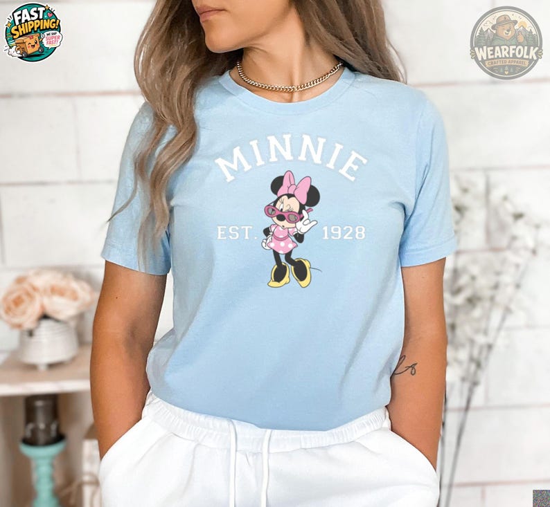 A woman wearing a light blue t-shirt featuring Minnie Mouse in sunglasses and a pink polka dot dress, with white text reading "MINNIE EST. 1928" on the chest.