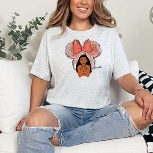 A woman wearing a light gray tee with an illustration of Moana's face framed by a pink mouse ear bow and the name "Moana" written in script.