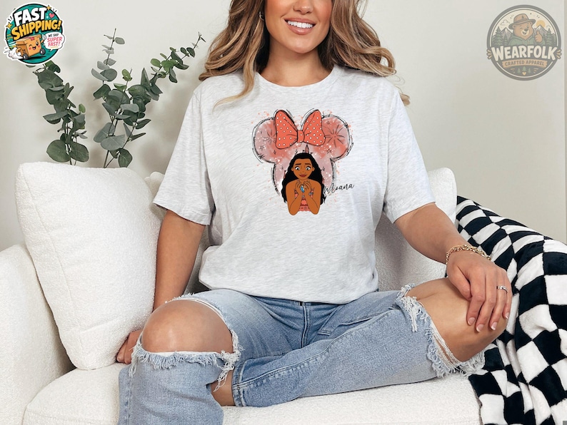 A woman wearing a light gray tee with an illustration of Moana's face framed by a pink mouse ear bow and the name "Moana" written in script.