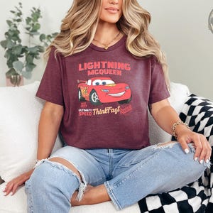 A woman wearing a heather maroon tee with a Lightning McQueen graphic and text that reads "Lightning McQueen est. 1977 Ultimate Speed ThunderFast Riviera".