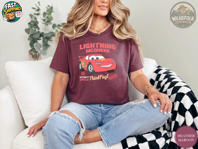 A woman wearing a heather maroon tee with a Lightning McQueen graphic and text that reads "Lightning McQueen est. 1977 Ultimate Speed ThunderFast Riviera".