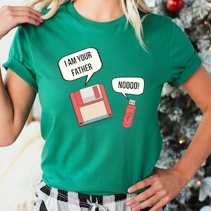Peut inclure: Un t-shirt vert kelly à manches courtes avec un motif représentant une disquette rouge et une clé USB rouge. La disquette a une bulle de texte indiquant I AM YOUR FATHER et la clé USB a une bulle de texte indiquant NOOOO!