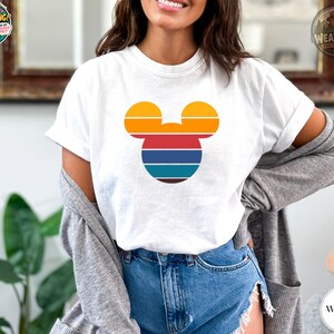 A woman wearing a white tee with a Mickey Mouse head graphic in orange, red, blue, and teal horizontal stripes.