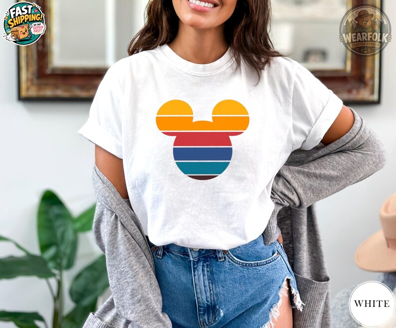 A woman wearing a white tee with a Mickey Mouse head graphic in orange, red, blue, and teal horizontal stripes.