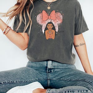 Woman wearing a charcoal gray Bella Canvas tee with a graphic of a girl with Mickey ears and a red polka dot bow surrounded by sparkles and text.