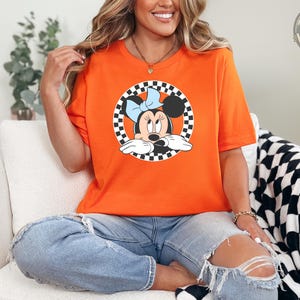 May include: Orange t-shirt featuring a Minnie Mouse graphic within a black and white checkered circle. The cartoon character has a blue bow. The shirt is made of a soft material and is suitable for casual wear.