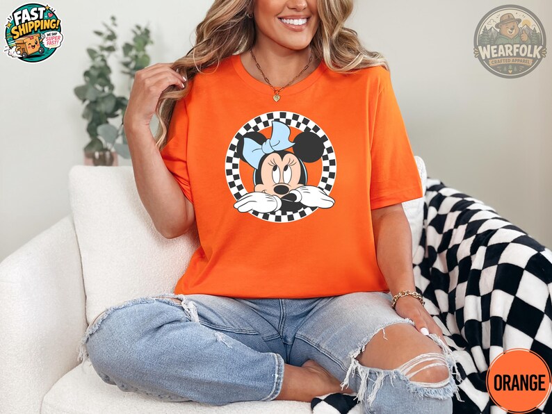 May include: Orange t-shirt featuring a Minnie Mouse graphic within a black and white checkered circle. The cartoon character has a blue bow. The shirt is made of a soft material and is suitable for casual wear.