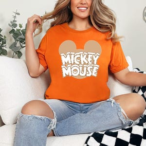 A woman wearing an orange tee with a Mickey Mouse graphic and white text that says "Mickey Mouse" centered on the chest.