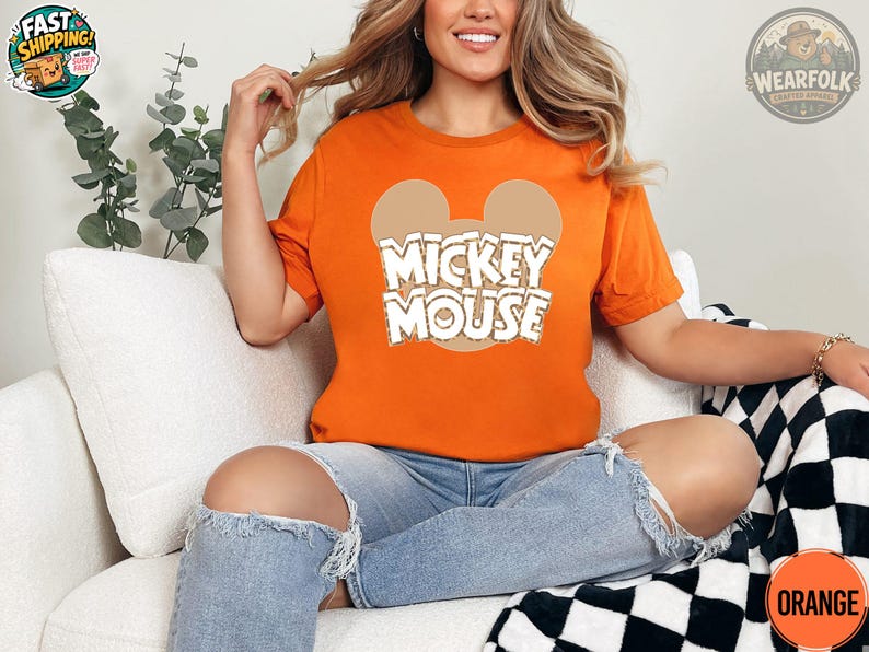A woman wearing an orange tee with a Mickey Mouse graphic and white text that says "Mickey Mouse" centered on the chest.