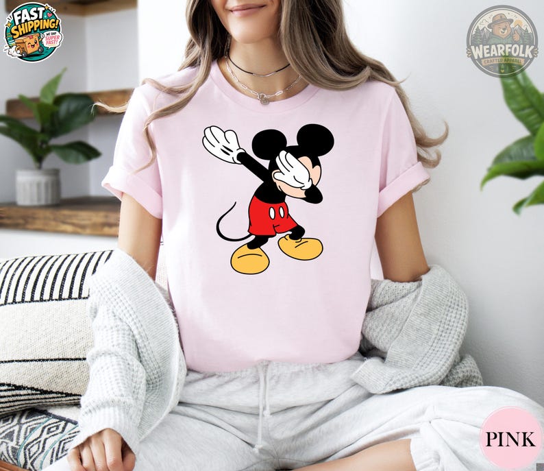 A woman wearing a light pink tee with a classic Mickey Mouse graphic showing the character with his arms raised wearing red shorts and yellow shoes.
