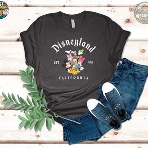 K&ouml;nnte beinhalten: Dunkelgraues T-Shirt mit Disneyland-Logo und Disney-Figuren. Der Text "EST 1955 CALIFORNIA" ist auf dem Shirt abgebildet. Das Bild zeigt auch blaue Jeans und Turnschuhe.