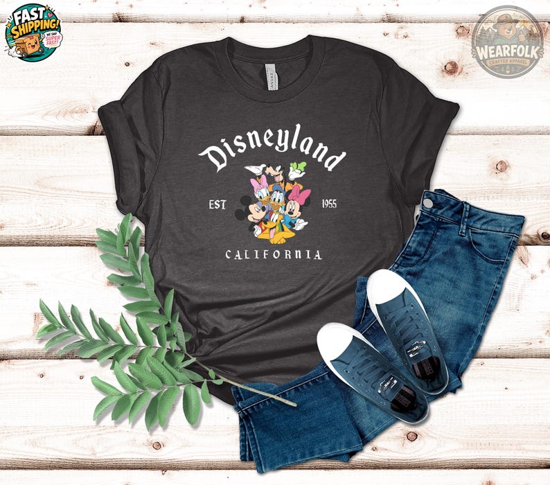 K&ouml;nnte beinhalten: Dunkelgraues T-Shirt mit Disneyland-Logo und Disney-Figuren. Der Text "EST 1955 CALIFORNIA" ist auf dem Shirt abgebildet. Das Bild zeigt auch blaue Jeans und Turnschuhe.