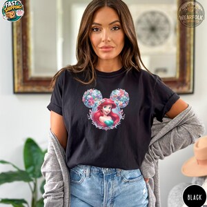 A woman wearing a black Bella Canvas tee with a Disney princess graphic featuring Ariel's face in the center of a floral Mickey Mouse ears design.