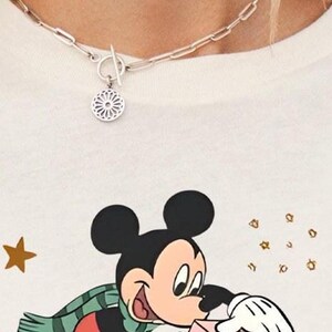 May include: A silver-toned chain necklace with a toggle clasp and a circular pendant featuring a geometric cutout pattern. Below the necklace is a white t-shirt with a graphic print of Mickey Mouse wearing a green scarf and holding a pink object.