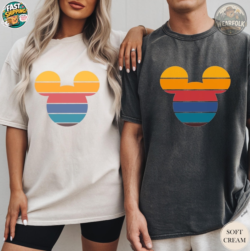 A woman in a cream tee and man in a charcoal tee wearing matching mouse head graphic tees with colorful horizontal stripes in yellow, red, blue, and teal.