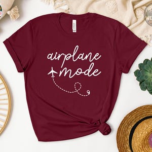 Flat lay of a heather maroon unisex t-shirt with white cursive text reading "airplane mode" and a small airplane graphic with a dashed flight path trail on the chest.