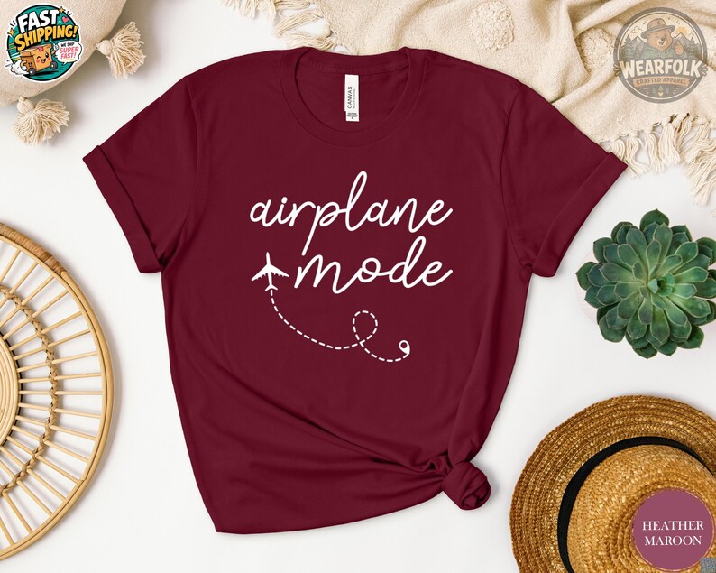 Flat lay of a heather maroon unisex t-shirt with white cursive text reading "airplane mode" and a small airplane graphic with a dashed flight path trail on the chest.