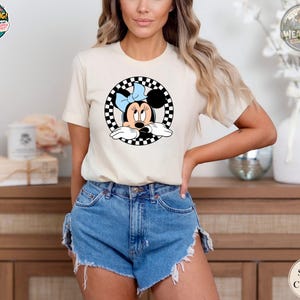 A woman wearing a cream Bella Canvas tee with a vintage Minnie Mouse graphic inside a black and white checkered circle design.