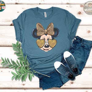 May include: Teal t-shirt featuring a Minnie Mouse graphic. Minnie is wearing heart-shaped gold glitter sunglasses and a leopard print bow. The shirt is paired with blue jeans and sneakers.