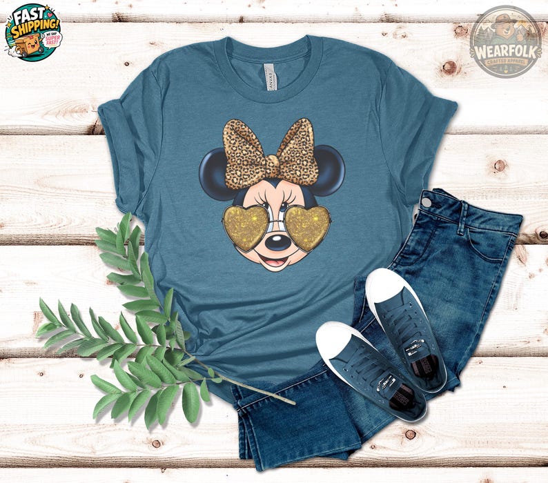 May include: Teal t-shirt featuring a Minnie Mouse graphic. Minnie is wearing heart-shaped gold glitter sunglasses and a leopard print bow. The shirt is paired with blue jeans and sneakers.