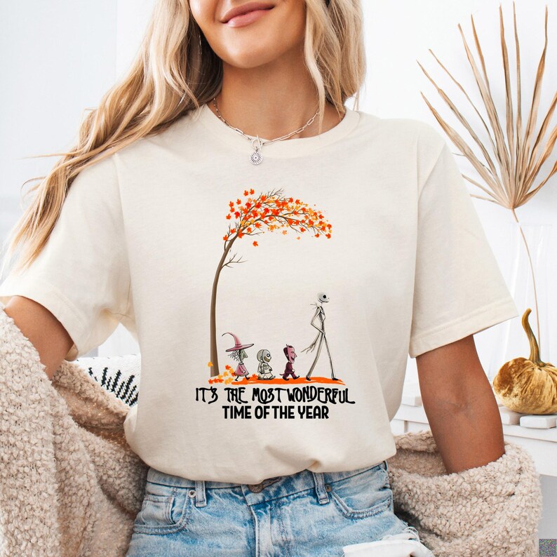 A woman wearing a cream Bella Canvas tee with a colorful autumn tree and Halloween characters below text reading "It's The Most Wonderful Time Of The Year".