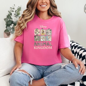 A woman wearing a pink t-shirt featuring Disney characters in safari outfits arranged in a grid, with text reading "Disney Animal Kingdom" in tan lettering.