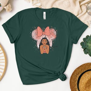 A woman wearing a dark green Bella Canvas tee with an illustration of a girl with long black hair below a pink bow and mouse ears graphic with sparkles.