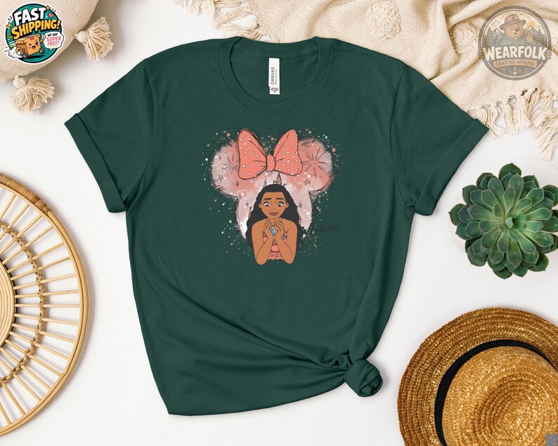 A woman wearing a dark green Bella Canvas tee with an illustration of a girl with long black hair below a pink bow and mouse ears graphic with sparkles.